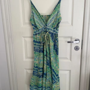 Grön och blå långklänning från Ethnix - En vacker långklänning från Ethnix med ett mönster i gröna och blåa nyanser. Klänningen har smala axelband och en knytning i midjan för en justerbar passform.