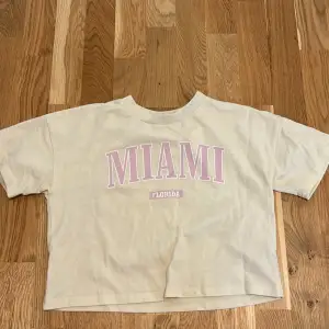 En gul t-shirt från Lindex med korta ärmar och texten 'MIAMI FLORIDA' i rosa på framsidan. T-shirten är lite croppad.