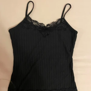 Svart linne med spetsdetaljer - Ett fint svart linne med smala axelband och spetsdetaljer vid halsringningen och nederkanten. Linnet har en ribbad struktur och är tillverkat i ett mjukt och bekvämt material. Aldrig använt. Skriv gärna om ni har frågor 💕