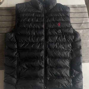 Ralph Lauren Väst - Säljer denna svarta ralph lauren dun västen då den ej används, Det är en lightweight väst och kan packas ner genom en inbyggd påse i fickan. Använd enstaka gånger. Den är i storlek M och köpt på Zalando. Orginal pris-3200kr
