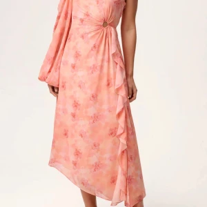 Ljusrosa blommig långklänning från Adoore - Säljer en superfin ljusrosa långklänning från Adoore med blommigt mönster och asymmetrisk design. Klänningen har oneshoulder, en lång puffärm och snygg volang längs sidan. Detalj med ring i midjan ger extra cool vibe. Perfekt för dig som vill sticka ut!