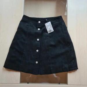 Svart jeanskjol med knappar - Snygg svart jeanskjol från H&M Divided i A-linjeformad modell. Kjolen har fem silvriga knappar framtill och är tillverkad i ett stadigt denimtyg. Perfekt att matcha med sneakers eller boots för en cool look.