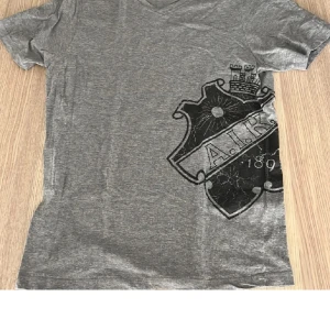 Grå AIK t-shirt med v-ringning - Snygg grå t-shirt från AIK med stort svart klubbmärke tryckt på framsidan. V-ringad hals och korta ärmar. Tillverkad i 100% bomull, perfekt för dig som vill visa ditt AIK-supporterhjärta. Officiell merchandise.