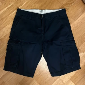 Svarta cargo shorts Dobber  - Snygga svarta cargo shorts från Ralph Lauren med klassisk design och praktiska fickor på sidorna. Sköna att bära med normal passform och tillverkade i mjuk bomull. Perfekta för sommardagar och chill häng utomhus.