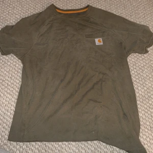 Olivgrön Carhartt t-shirt med ficka - Snygg olivgrön t-shirt från Carhartt med klassisk logga på bröstfickan. Modellen är enkel med rund hals och korta ärmar, perfekt för en chill och avslappnad stil. Materialet är mjuk bomull som känns skönt mot huden.
