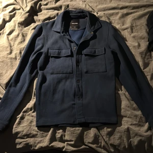 Overshirt från Jack & Jones - Snygg mörkblå overshirt från Jack & Jones med klassisk krage och två stora bröstfickor med lock. Jackan har knappar framtill och är tillverkad i ett mjukt, strukturerat material som känns både stilrent och avslappnat.