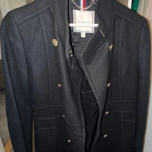 Svart rock från Tommy Hilfiger - Stilren svart rock från Tommy Hilfiger med snygga guldknappar och markerade axlar. Jackan har dubbelknäppning och två fickor framtill. Perfekt för dig som gillar klassisk och tidlös design med en modern twist.