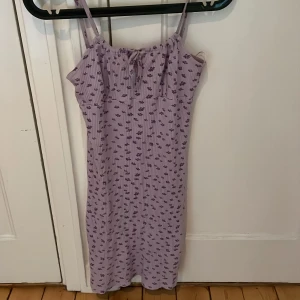 Ljuslila blommig klänning Stories XS - Supersöt ljuslila kortklänning från Stories i storlek XS. Klänningen har smala axelband, ribbad struktur och ett litet knyt framtill. Blommigt mönster i lila över hela klänningen ger en fräsch vibe. Perfekt för varma dagar och sommarhäng.