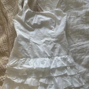 Vit kort klänning med volanger - Säljer en söt vit kort klänning med volanger nedtill och breda axelband. Klänningen har en enkel och fräsch design som ger en romantisk vibe. Perfekt för varma dagar och passar dig som gillar en feminin stil.