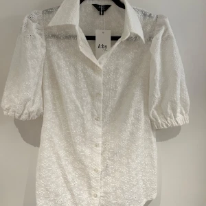 Vit broderad skjorta från A:by Anna - Superfin vit skjorta från A:by Anna med broderade blommor och puffiga korta ärmar. Skjortan har klassisk krage, knappar framtill och är gjord i bomull. Perfekt för dig som gillar romantiska detaljer och en luftig känsla.
