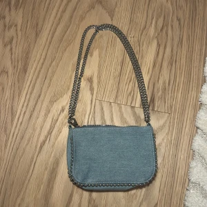 Ljusblå axelväska i jeans med kedja - Snygg ljusblå axelväska från H&M i jeansmaterial med silverfärgad kedjerem och coola nitar längs kanterna. Väskan har dragkedja upptill och är perfekt för att bära det viktigaste. Enkel men trendig design som lyfter vilken outfit som helst.