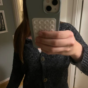 Glittrig svart kofta med stora knappar - Säljer en svart glittrig kofta med stora silvriga knappar framtill. Koftan är stickad och har en croppad passform med långa ärmar och ribbade muddar. Perfekt för dig som vill ha något extra till outfiten.