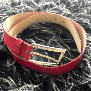 Rött bälte/ skärp H&M- strass-spänne Made in Italy - Ett fint rött bälte/ skärp från H&M med ett elegant, rektangulärt spänne dekorerat med swarowskistenar. En speciell detalj är att spännet är stämplat med ”Made in Italy”, vilket tyder på att det kan komma från någon av H&Ms premiumkollektioner. Det är i ett fint begagnat skick med spår av användning, se foto. Några av swarowskistenarna på spännet har mörknat eller lossnat, men det ger bara bältet karaktär och det är fortfarande mycket effektfullt.  Mått: L 89 cm B 3 cm