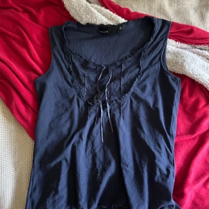 Mörkblå ärmlös topp från KappAhl - Snygg mörkblå ärmlös topp från KappAhl i storlek M (40/42). Toppen har en djup rundad ringning med spetsdetaljer och en söt knytning framtill. Perfekt att matcha med jeans eller kjol för en chill look.