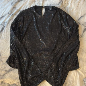 Svart paljettblus från Amisu - Glittrig svart blus från Amisu i storlek S, täckt med paljetter som ger en riktigt festlig vibe. Blusen har rund hals och långa ärmar, perfekt för att sticka ut på nästa party eller event. Snyggt nyckelhål bak i nacken för extra detalj.