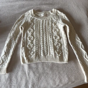 Vit stickad tröja Holly & Whyte - Säljer en vit stickad tröja från Holly & Whyte med snygga kabelstickade detaljer och pärlor på framsidan. Tröjan har rund hals, långärmade ärmar och ribbade muddar. Perfekt för dig som gillar klassisk stil med en twist.