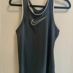 Svart linnetopp från Nike - Sportig svart linnetopp från Nike med vitt kantband och klassisk vit swoosh-logga framtill. Perfekt för träning med sitt svala material och ärmlösa design. Snygg och enkel att matcha med träningskläder.