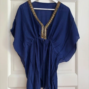 Marinblå kaftanklänning med paljetter - Snygg marinblå kaftanklänning med v-ringad hals och glittrande paljettdekor i guld längs halsringningen. Klänningen har ett löst och luftigt fall, knyt i midjan och är tillverkad i ett tunt, skönt material. Perfekt för dig som gillar boho-vibe och vill sticka ut.