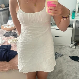Vit kort sommarklänning från Bershka - Supersöt vit klänning från Bershka med smala axelband och lätt rynkad byst. Modellen är kort och har en enkel, flowy passform som ger en chill vibe. Perfekt för varma dagar och enkel att styla med smycken eller sneakers. Materialet är mjukt och känns skönt mot huden. Har bara använts 2 gånger. 