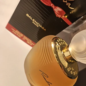 Rumba Eau de Toilette Balenciaga - Rumba från Balenciaga är en elegant parfym i en lyxig, bärnstensfärgad flaska med gulddetaljer och en frostad, rund kork. Förpackningen är svart med röda och guldiga detaljer och en röd rosett. Doften är klassisk och tidlös, perfekt för dig som gillar exklusiva parfymer.VINTAGE