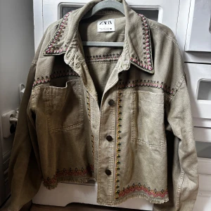 Broderad jeansjacka från Zara, S - Beige jeansjacka från Zara med broderade detaljer i rosa, svart och gult längs krage, bröst och nederkant. Jackan har knappar framtill, två stora fickor och fransad nederkant. 