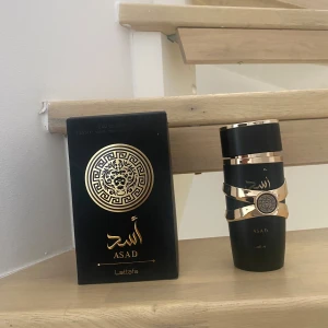 Lattafa Asad Eau de Parfum 100ml - Lattafa Asad Eau de Parfum i en lyxig svart flaska med guldiga detaljer och ett lejonmotiv. Flaskan har en unik form med guldfärgade band runtom och arabiska inskriptioner. Kommer i en elegant svart och guldig kartong. Volym: 100 ml. Men har användit lite så det är 90ml kvar.