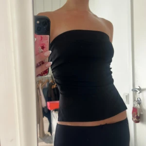 Svart bandeau tubtopp med slits - Säljer en svart bandeau tubtopp med tight passform och snygg slits på sidan. Toppen är axelbandslös och sitter smickrande mot kroppen, perfekt för dig som gillar minimalistisk stil. Enkel att matcha med både jeans och kjol.