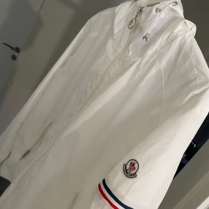 Vit vindjacka från Moncler med huva - Snygg vit vindjacka från Moncler med huva och dragkedja framtill. Jackan har Moncler-logga på ärmen och coola ränder i rött och blått vid ärmslutet. Praktiska fickor med dragkedja och justerbar snörning i huvan. Perfekt för dig som gillar stilren och sportig look.