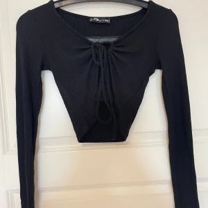 Svart croppad ribbad topp med knyt - Svart långärmad croppad i ribbad bomull. Toppen har snygg knytning framtill och en lätt v-ringad hals. Perfekt att styla med jeans eller kjol för en trendig look.