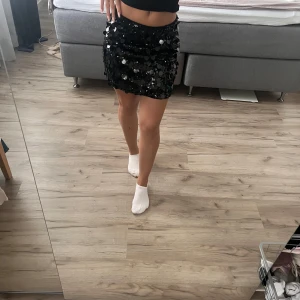 Svart paljettkjol med glittriga detaljer - Säljer en svart kortkjol täckt med stora glittriga paljetter som ger en riktigt festlig vibe. Kjolen har rak form och sitter lågt i midjan. Perfekt för dig som vill sticka ut och glänsa på nästa event!🌺