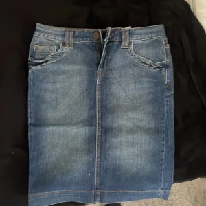 Blå jeanskjol med fickor och slits - Säljer en klassisk blå jeanskjol med fem fickor, bälteshällor och snygga kontrastsömmar. Kjolen har dragkedja och knapp framtill samt en slits bak för extra rörelsefrihet. Perfekt för dig som gillar denim och vill ha en enkel, stilren look.