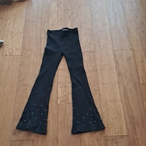 Svarta bootcut byxor från Kappahl - Svarta ribbade bootcut byxor från Kappahl i stretchigt material. Byxorna har utsvängda ben med små pärldetaljer längst ner för en extra touch. Perfekta för dig som gillar en trendig och bekväm stil. 