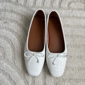 Vita ballerinaskor med rosett från NA-KD - Snygga vita ballerinaskor från NA-KD med flätad struktur och söt rosettdetalj framtill. Fyrkantig tå och mjuk sula. Använda en gång så de är som nya. Säljer då de tyvärr är förstora. Ordinarie pris är 499 kr. 