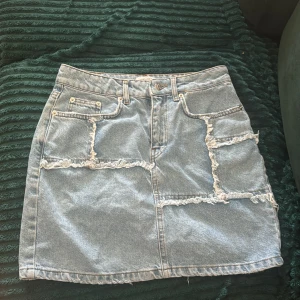 Patchad jeanskjol från NA-KD - Säljer en ljusblå jeanskjol från NA-KD med patchade detaljer och råa, fransiga sömmar. Kjolen har klassisk femficksdesign, bälteshällor och knappstängning framtill. Perfekt för dig som gillar en edgy och trendig stil.