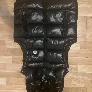 Svart dunväst från Moncler med huva - Säljer en svart, glansig dunväst från Moncler i storlek S. Västen har två dragkedjefickor framtill, huva och klassisk Moncler-logga på bröstet. Perfekt för lager-på-lager och riktigt snygg till streetwear-stilen.