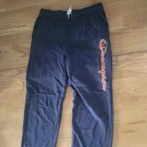 Mörkblå mjukisbyxor från Champion - Mörkblå mjukisbyxor från Champion med stort logotryck längs ena benet i rött och vitt. Byxorna har resår och snörning i midjan samt muddar vid bensluten. Perfekta för chill eller träning, riktigt sköna och stilrena.