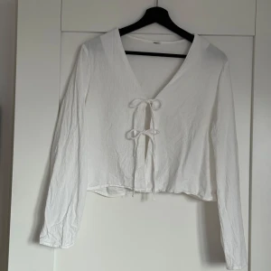 Vit långärmad blus med knyt - Säljer en vit blus med långa ärmar och två söta knyt framtill. Toppen har v-ringning och är lätt croppad, perfekt att styla med jeans eller kjol. Tunt och luftigt material som känns skönt mot huden.