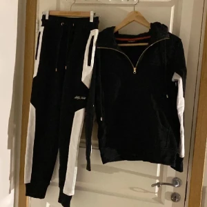 Hoddie och byxor  från Elly Pistol sett  - Svart hoodie från Elly Pistol i storlek S med vita detaljer längs ärmarna och stor vit logga på ryggen. Tröjan har huva, half zip-dragkedja och snörning vid halsen. Materialet är mjukt och känns som bomull, perfekt för chill eller träning. Båda delarna o storlek S 
