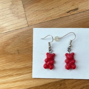 Röda örhängen med nallebjörnar - Säljer ett par söta örhängen med röda nallebjörnar i plast. Hängande modell med silvriga krokar som ger en lekfull och unik touch till din stil. Perfekta för dig som gillar roliga accessoarer och vill sticka ut lite extra. Jag själv är nickelallergiker och har inte haft några problem. 