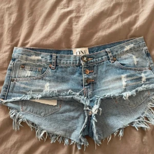 Blå jeansshorts med fransar från One - Snygga blå jeansshorts från One Teaspoon med slitna detaljer och råa fransade kanter. Modellen har hög midja, tre knappar framtill och klassiska fickor både fram och bak. Perfekt för dig som gillar en edgy och avslappnad stil. Passar storlek M men är storlek 31 i midjan.