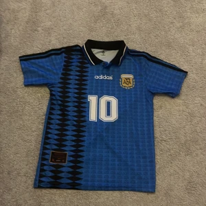 Argentina retro fotbollströja Adidas M - Retro Argentina fotbollströja från Adidas i blått med svarta detaljer och tryckt nummer 10. Tröjan har korta ärmar, pikékrage med knapp och AFA-emblem på bröstet. Tillverkad i lätt och ventilerande ClimaCool-material. Snyggt geometriskt mönster längs ena sidan.