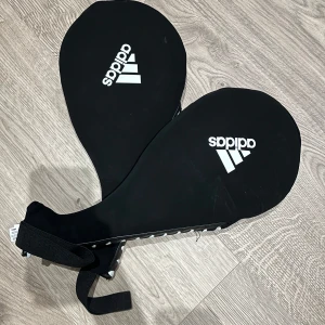 Sparkmitts - Bilden visar två svarta Adidas sparkmittsar, perfekta för träning inom kampsport eller fitness. De har tydlig Adidas-logga och justerbara remmar för bra passform. Perfekt för dig som vill förbättra din teknik och styrka på ett roligt sätt.