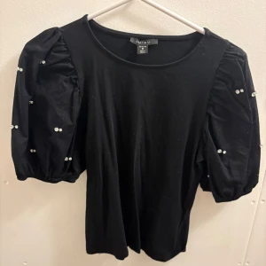 Svart topp med puffärmar och pärlor  - Svart topp från New Yorker med puffiga kortärmar dekorerade med glittrande pärlor. Toppens kropp är i mjuk bomullskänsla och ärmarna har en ballongformad look som ger extra volym. Perfekt för dig som vill sticka ut med coola detaljer. Aldrig använt 