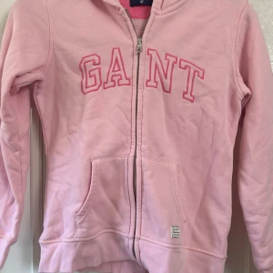 Rosa hoodie från GANT med dragkedja - Säljer en ljusrosa hoodie från GANT med broderad logga i rosa på bröstet. Tröjan har huva, långa ärmar och praktiska fickor framtill. Materialet är mjuk bomull och den har en dragkedja hela vägen. Perfekt för chill dagar och snygg till jeans.