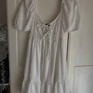 Vit kort klänning från Gina Tricot - Supersöt vit klänning från Gina Tricot i storlek S. Den har puffiga korta ärmar, knytning framtill och en volangkant nedtill. Materialet är mjukt och strukturerat, perfekt för varma dagar. ❣️❣️❣️