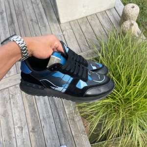Valentino Rockrunner sneakers blå/svart - Snygga Valentino Rockrunner sneakers med blått och svart camouflagemönster. Skorna har svarta mockadetaljer, svarta snören och coola nitar på sulan. Materialmix av skinn och mocka ger en lyxig känsla. Perfekta för dig som vill sticka ut med stil. Du får med box och dustbags och exktra skosnören 