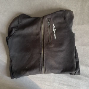 Svart hoodie från Sail Racing - Svart hoodie från Sail Racing med hel dragkedja och huva. Tröjan har en bröstficka med dragkedja och logotypen tryckt på vänster sida. Mjukt material och ribbade muddar vid ärmslut och nederkant. Perfekt för chill dagar eller när du vill ha en sportig look.