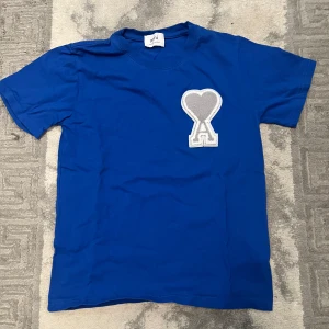 Blå Ami t-shirt med hjärtdetalj - Snygg blå t-shirt från Ami med korta ärmar och ett stort grått hjärta med bokstaven A broderat på bröstet. Rund hals och mjukt bomullsmaterial gör den skön att bära. Perfekt för dig som gillar stilrena plagg med en twist.