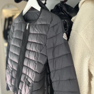 Svart Moncler cardigan - Snygg svart cardigan/dunjacka från Moncler med quiltad design och rund halsringning. Jackan har dragkedja framtill, två fickor med svarta detaljer och klassisk Moncler-logga på ärmen. Perfekt för dig som vill ha en lätt och stilren jacka till höst och vinter. Köpt förra året nästan helt oanvänd direkt från moncler butiken från birgerjarlsgatan, nypris 9900kr💞