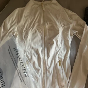 Vit define jacka från Lululemon - Snygg svart träningsjacka från Lululemon med guldiga dragkedjor och figurnära passform. Jackan har hög krage, långa ärmar och två fickor med dragkedja. Jackan är oanvänd och lappen sitter kvar i jackan. Lululemon dustbag följer med.